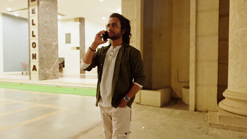 Man talking on mobile phone in a building hallway, Young person on call in lobby, Men walk and talk alone, with long hair and a beard, Columns and marble flooring, is calling someone, Indian, 20s.
