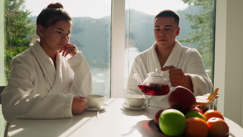 Man pours tea into cup sitting with wife at table. Young couple spends morning drinking hot tea together in kitchen. Concept of warm family relationship