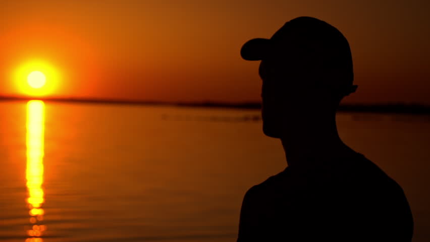 A man stands in silhouette, sipping from a cup as he gazes out over the tranquil lake at sunset. The vibrant orange sky creates a breathtaking backdrop for a moment of peaceful reflection.