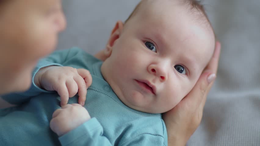 Mom playing with a newborn baby - slow motion. A cute child is trying to pronounce sounds while looking at his mom. The face of an active caucasian infant looking at a parent. Happy male kid is lookin