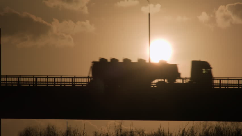 Silhouette of fuel truck with gasoline tanker driving on road bridge on background of sunset sky in evening time.