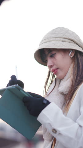 Asian woman listening to the music on earphones and using digital pen on tablet by the river in Tokyo city, Japan. Attractive girl freelance work anywhere with using gadget device and online network.