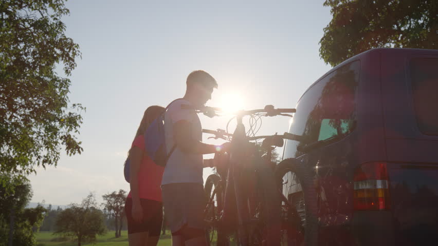 Young man and a woman preparing for off-road biking, taking down electric mountain bikes from the bike rack on the camper van, handheld shot.