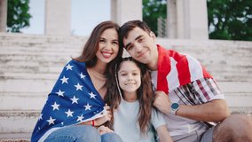 Portrait of A young family with a child girl is sitting on the steps and wrapped in the flag of the United States of America. Family celebrates American Independence Day. Group of people with USA flag - Powered by Shutterstock - Get 15% off with code: PIKWIZARD15