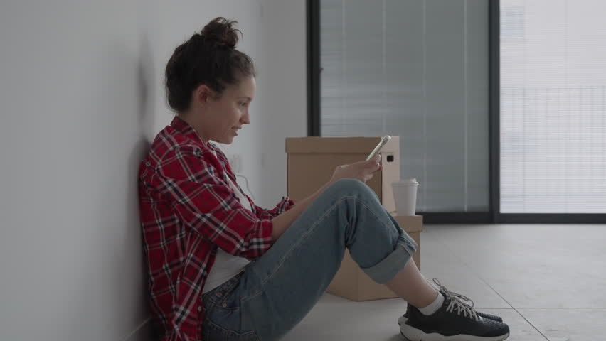 Profile shot of young woman sitting on apartment floor with boxes and texting. Holding smartphone mobile and typing while smiling and happy. Just moved into new home or moving out. Boxes on the floor