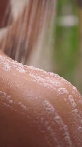 Vertical. Black woman cleaning foam skin with water relaxing in shower from neck to shoulder. Close-up unrecognizable young African female removing soap for daily hygiene. Personal body care in home. 