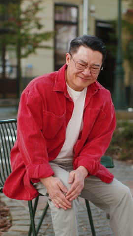 Sick Asian middle-aged man suffer from painful discomfort severe knee joint ache, leg muscles, could not walk outdoors. Chinese sad guy massaging knees illness cramps rheumatism on urban city street