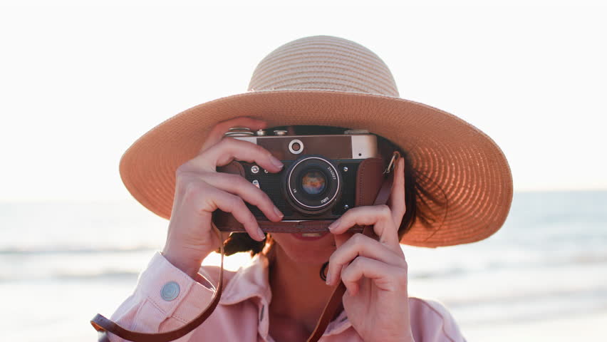 Beautiful woman in a hat takes pictures outdoors using an analog camera. a girl with a camera walks on the beach. travel and vacation.