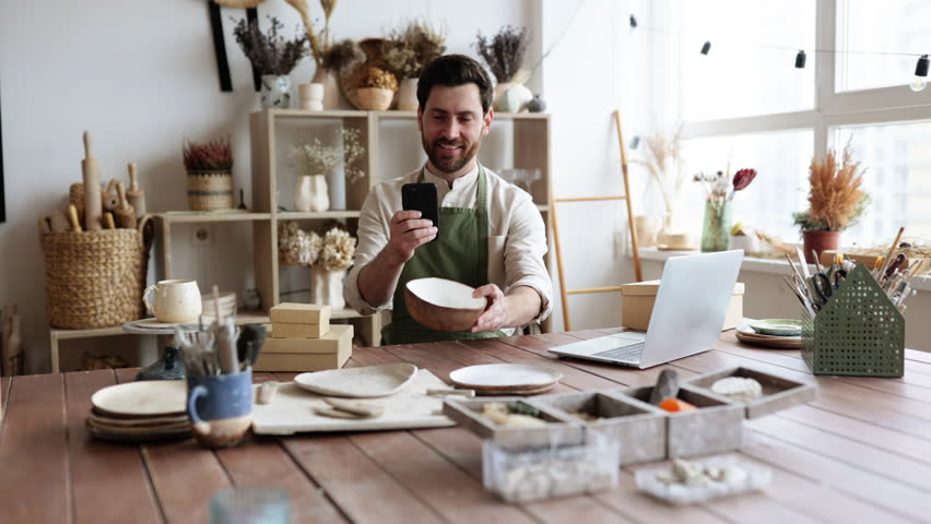 Charismatic Caucasian man seller in souvenir shop takes pictures of goods at phone for posting on social networks. Successful guy store employee with ceramic tableware shoots photos on smartphone.
