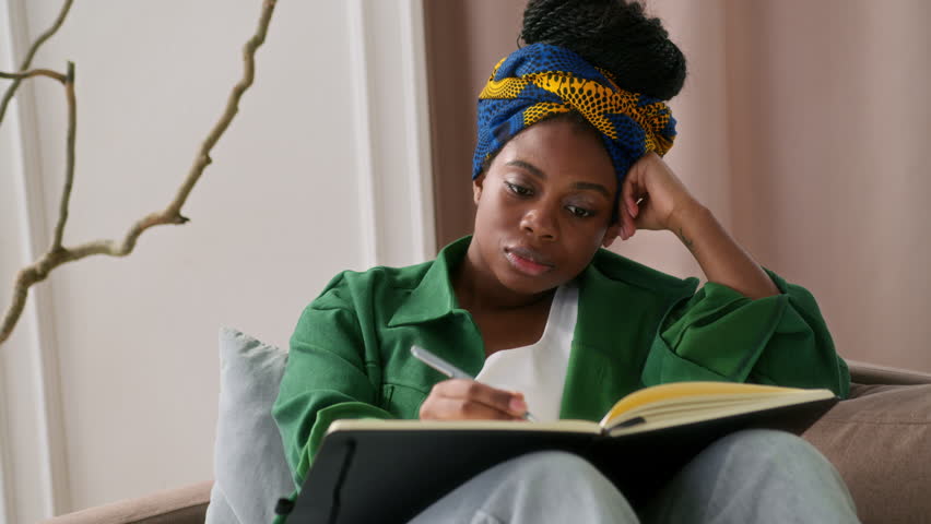 African American girl sits on couch alone, holding notebook and writing thoughts down in diary. Female black student taking notes or young writer working on a new story. She ponders and thinks over.
