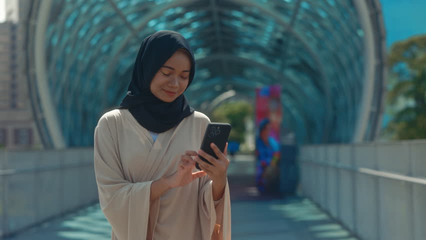 Beautiful Muslim Woman Standing Outside Near Modern Building Using her Smartphone. Young Girl Wearing Hijab Using Cell Phone and Surfing on the Internet. People and Technology
