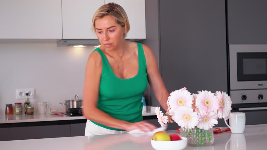 Happy woman cleaning kitchen island with friend placing empty tray on it. Smiling females are talking while preparing for lunch at home.