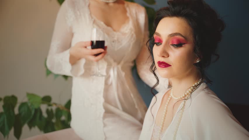 Two Fantasy women Sexy girl maid of honor beauty face red lips dark hair queen sitting on throne chair in luxury blue room. vintage silk white dress fairy style noble lady in waiting holds glass wine.