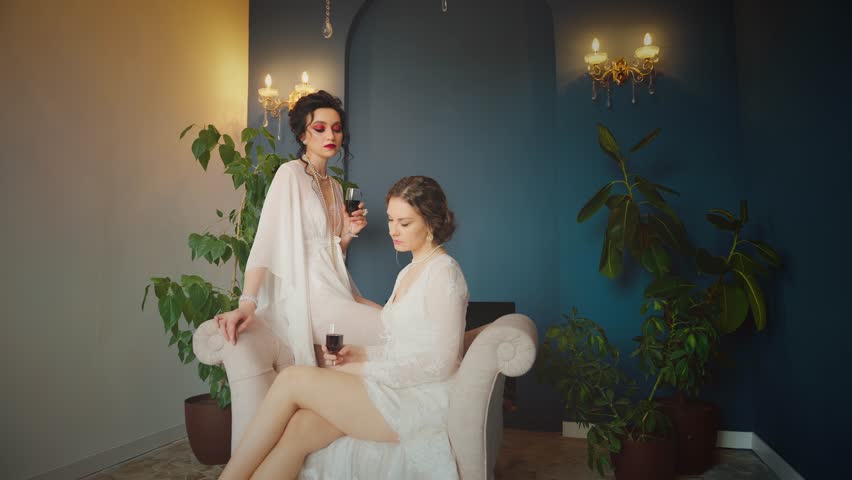 Two Fantasy women Sexy girl maid of honor beauty face red lips dark hair queen sitting on throne chair in luxury blue dark room. vintage silk white dress fairy style noble lady holds glass wine posing