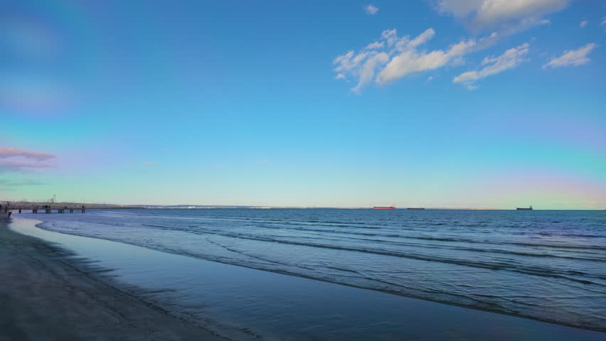 A serene beach scene with gentle waves lapping against the shore under a clear blue sky. Distant ships are visible on the horizon, enhancing the tranquil coastal atmosphere
