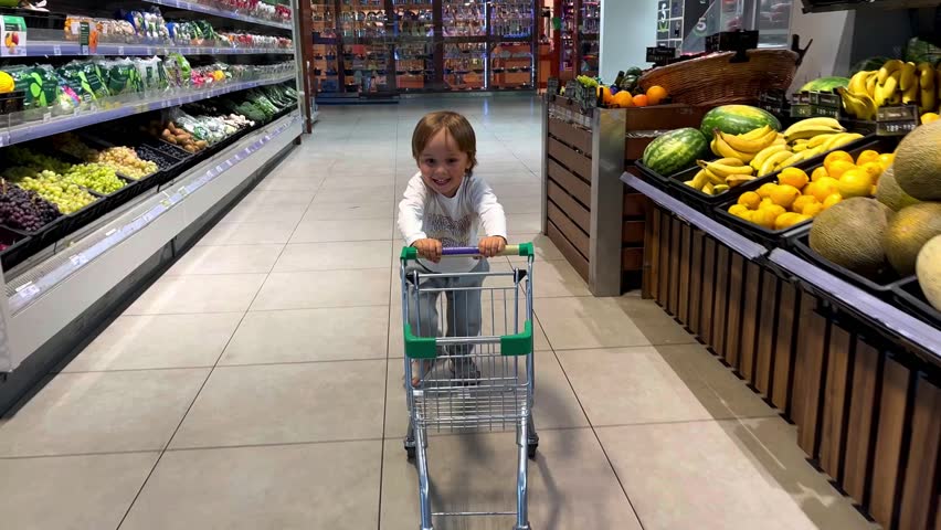 Adorable child is having fun running around the store with a cart past vegetables and fruits. The child is shopping at the supermarket. The boy buys healthy food. A playful kid customer in a store