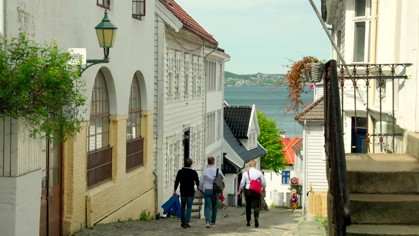 The beautiful alley of Skuteviksveien in Bergen, Norway. Old houses on both sides of the street, you can see the fjord in the background. Three persons walking away from the camera.