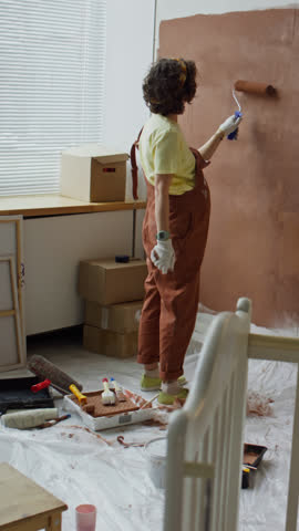 Vertical back full footage of single mother in late pregnancy stage painting walls with roller in brown while remodeling room into nursery for future child