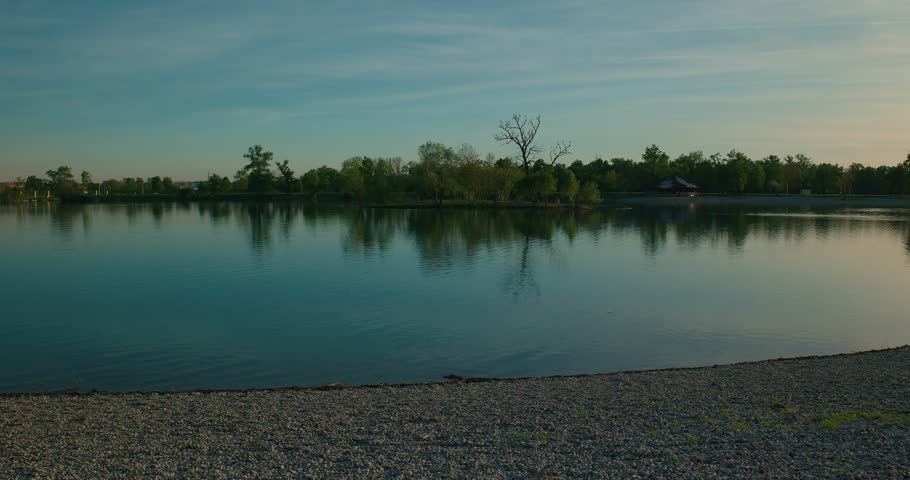 Calm lake with tree reflections, pebble shore, and a clear sky at Jarun Lake, Zagreb Croatia