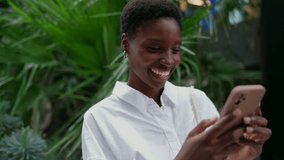 cheerful young Black woman joyfully engages with smartphone app outdoors. Female blogger smiles brightly while chatting online, enjoying a moment of connection and happiness with mobile phone - Powered by Shutterstock - Get 15% off with code: PIKWIZARD15