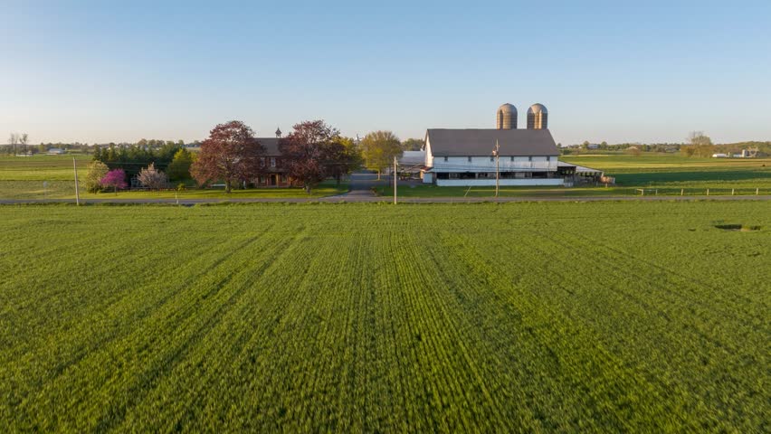 Golden hour sunset over classic American farm. House, barns and silos. Aerial hyperlapse, time lapse of traffic in front of countryside farmland.