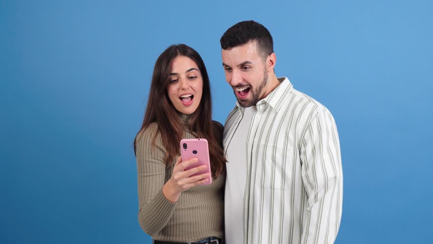 Caucasian couple having fun using a mobile phone to browse on internet with an application at blue studio background. Two friends betting online with a cellphone app on a turquoise colour backdrop