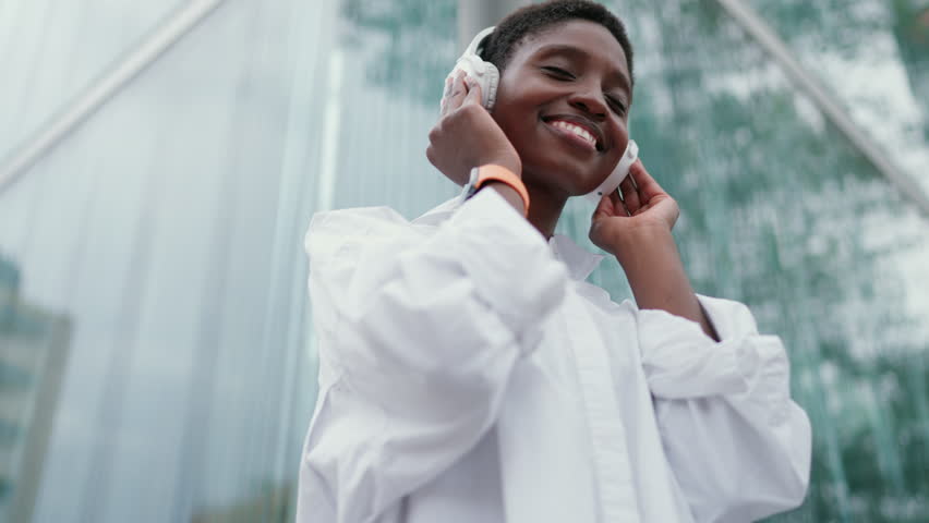 Confident woman with wireless headphones, immersed in music, feeling beat while dancing and singing outdoors in city. Female embodies vibrancy and energy of urban life and joy that comes from music