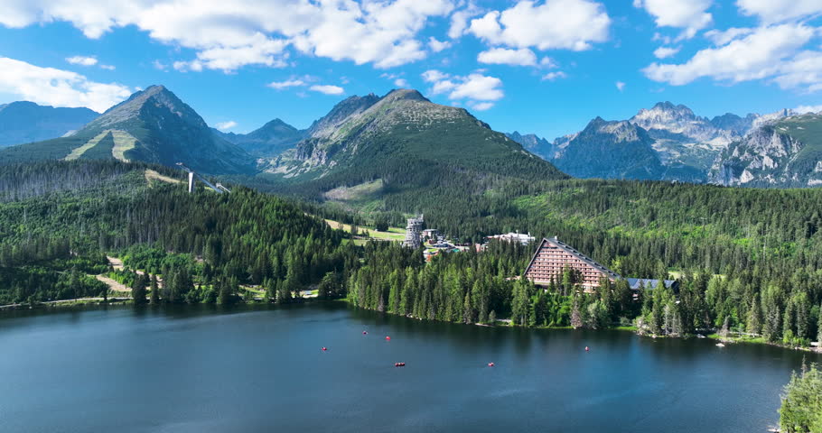 Drone footage of Štrbské Pleso in the High Tatras Mountains, Slovakia, showcases clear lake water, majestic peaks, pristine nature, and tranquil surroundings. Enjoy breathtaking views under blue skies