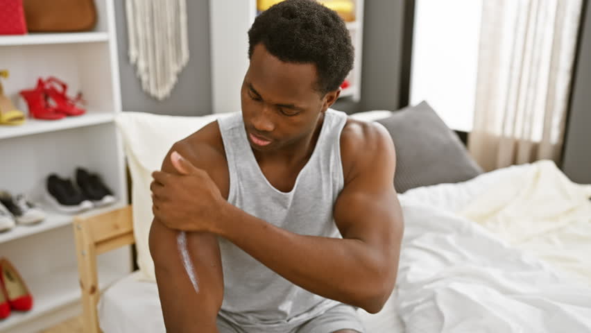 A young man applies lotion to his elbow in a well-lit modern bedroom.