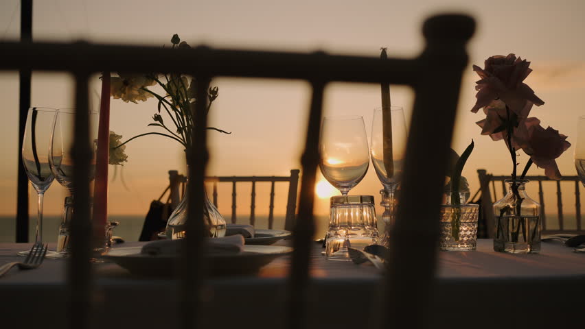 Romantic sunset dinner table honeymoon beach. Set up glasses with candles flowers for couple dinner party.