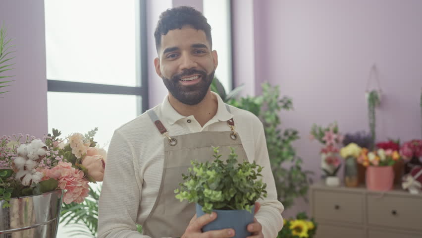 A smiling young man with a beard enjoys smelling a potted plant inside a bright, colorful flower shop.