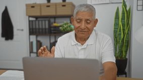 Mature hispanic man in a white shirt having a video call in an office setting with shelves and plants in the background.