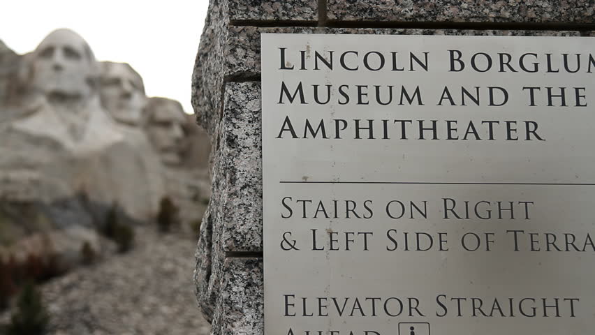 Mount Rushmore in the Distance: Signpost in Focus on a Cloudy Day. Mount Rushmore appears blurred in the background, with a signpost in the foreground on a cloudy day.