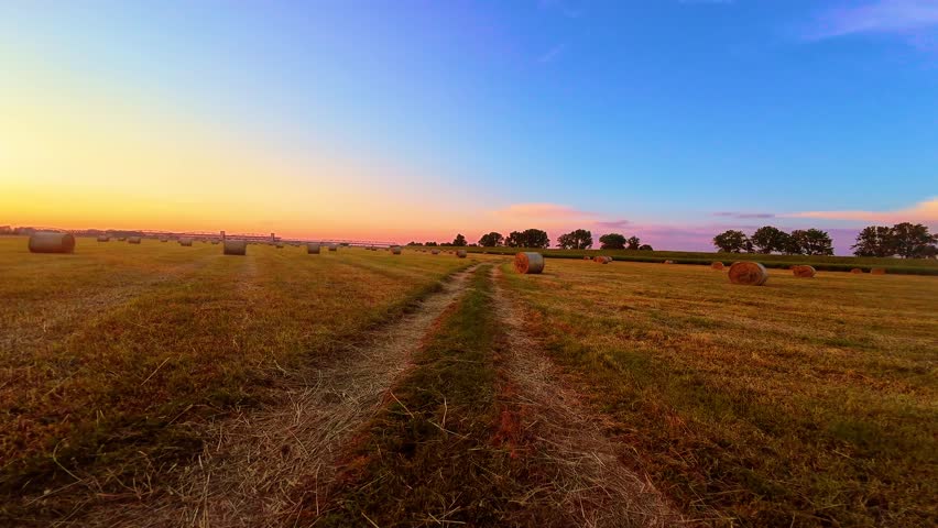 Hay Bales in Field at Sunset. POV tractor driving through rural countryside. Beautiful sunset over field with hay bales. Dirt road runs through middle of agricultural field