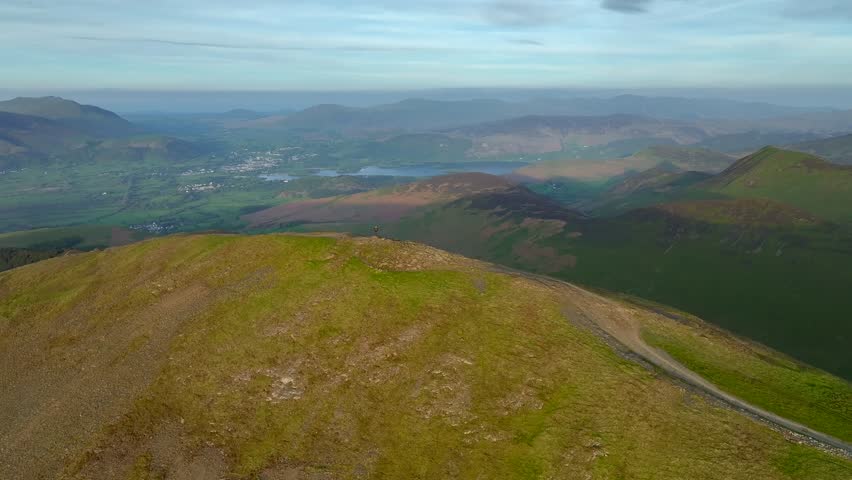 Distant lone hiker on rounded mountain summit at golden hour. Camera orbit expanding wide green valley and revealing Skiddaw mountain range. Grisedale Pike, Lake District, Cumbria, England, UK