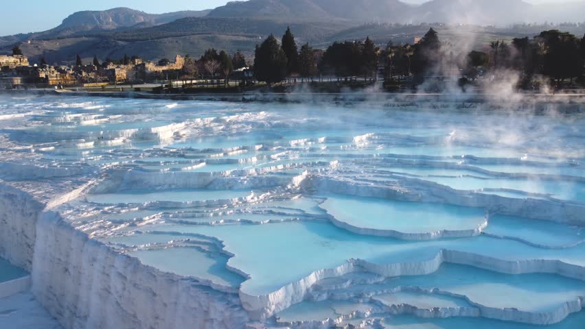 Steamy blue hot springs, mineral water, forming travertine limestone mineral deposit formations. Pamukkale, Turkey (Türkiye) UNESCO World Heritage Site. Looking toward ancient city of Hierapolis ruins