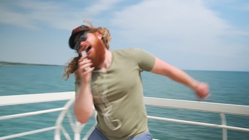 Slow motion, young caucasian redhead man sing swirl in sunglasses on wooden pier on seashore, positive hipster celebrating holiday. 