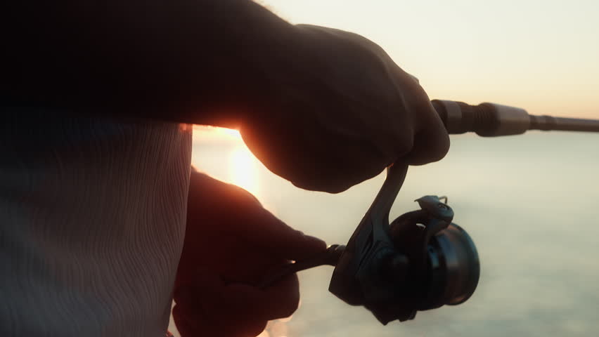 Close-up of a fisherman spins the reel on a spinning the fishing rod against of sunset