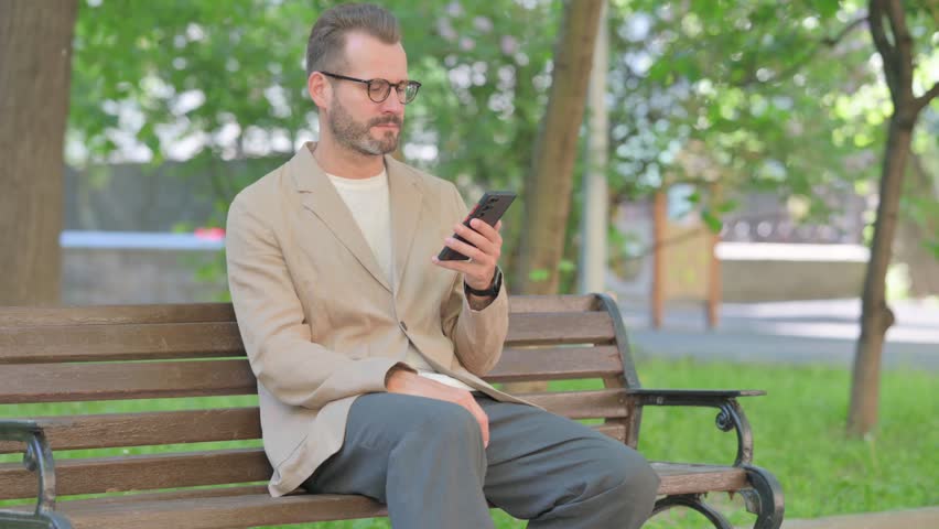Angry Middle Aged Man Talking on Phone in Argument while Sitting Outdoor on Bench