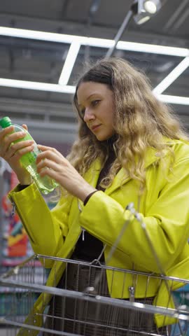 Woman checks ingredients list of soda drink and puts bottle back on rack. Female customer shocked by dangerous elements of beverage in supermarket. Vertical shot