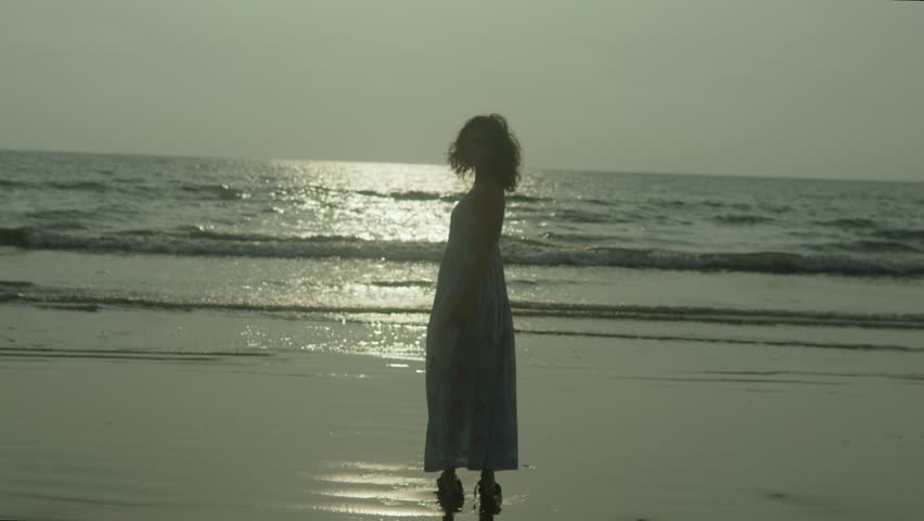 Indian woman silhouette stands on the beach in a white dress. The sky is cloudy and the sun is setting in India