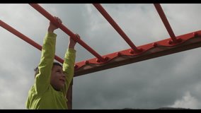 Child in a green jacket playing on monkey bars in an outdoor playground, with mountains and cloudy skies in the background, The child is engaged in outdoor sports - Powered by Shutterstock - Get 15% off with code: PIKWIZARD15