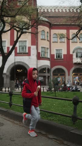 A joyful boy in a red hoodie jumps while holding an ice cream cone, set against an urban park backdrop with historic buildings