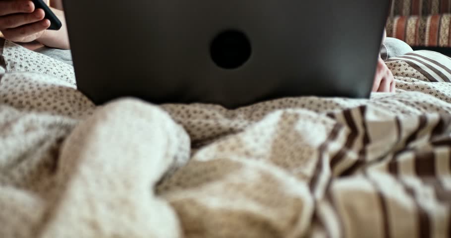 A young adult man sits on a mattress on the floor and enthusiastically works on a laptop or smartphone. The camera moves from bottom to top