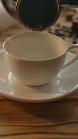 Pouring coffee from the cezve into a cup on a wooden table. Jute bag, oat liver and coffee beans. A cozy atmosphere for breakfast. Slow motion. Vertical shot.