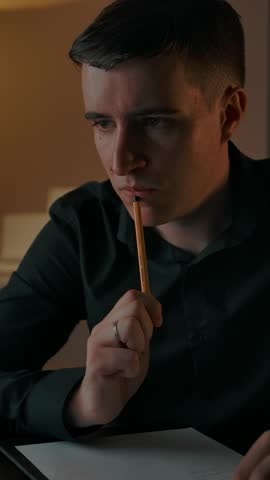 Caucasian male thinks about solving hard mathematic equation in online studying. Handsome student writes notes in text pad and look at laptop screen in night mood interior. Vertical shot.