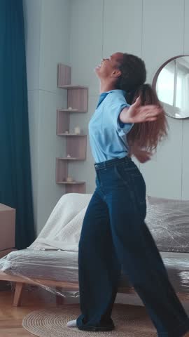 Young beautiful casual African American woman moved into own home performs festive dance rejoicing at availability of money for renting property located in living room with stylish interior.