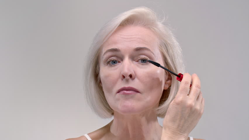 Old woman paints her eyelashes with black mascara. Senior female uses cosmetics at retirement age, is going on a date. Elderly lady takes care of the body and face, dry facial skin with wrinkles