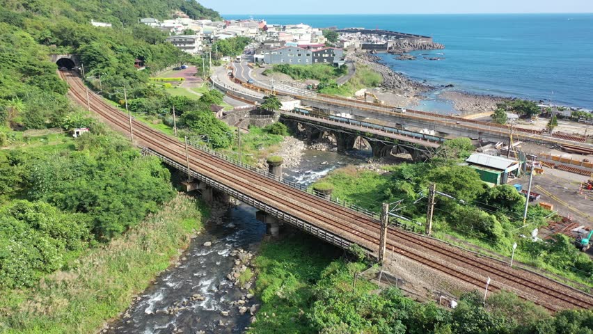 Aerial view of a fast train dashing across a river by the mountainside and a coastal highway running thru a seaside village next to Daxi Harbor on a sunny summer day, in Toucheng, Yilan, Taiwan