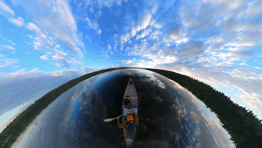 Man paddling a canoe on a very calm northern lake in the wide angle 360 degree type view. The calm water reflects the blue sky that has scattered white clouds. The sound of the paddling and birds are 
